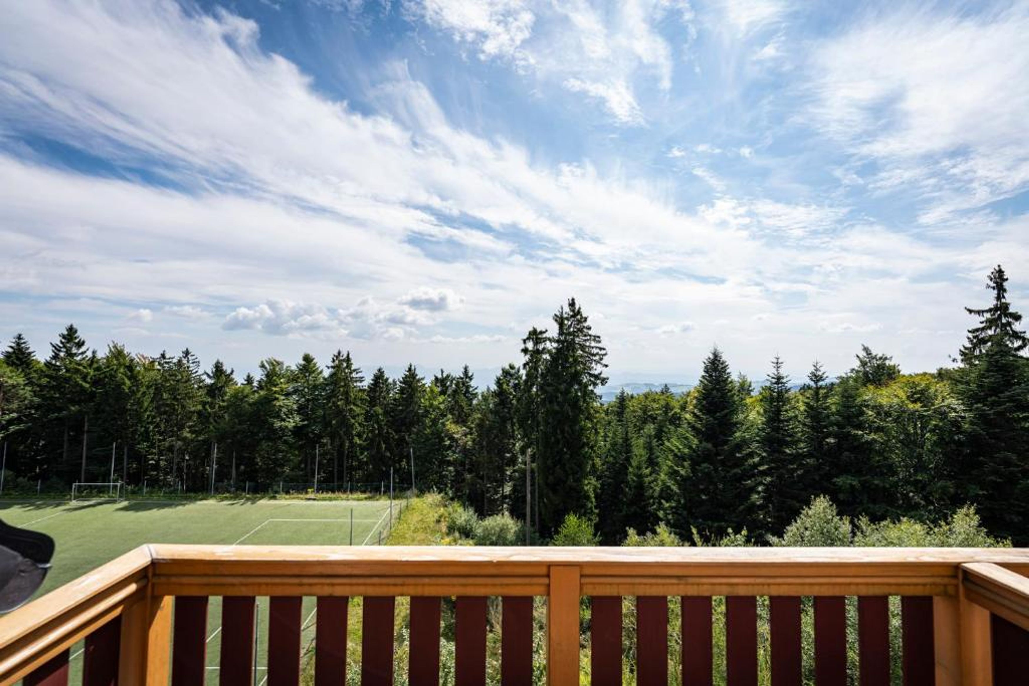 A tennis court with a wooden deck in the foreground and a forested area in the background.