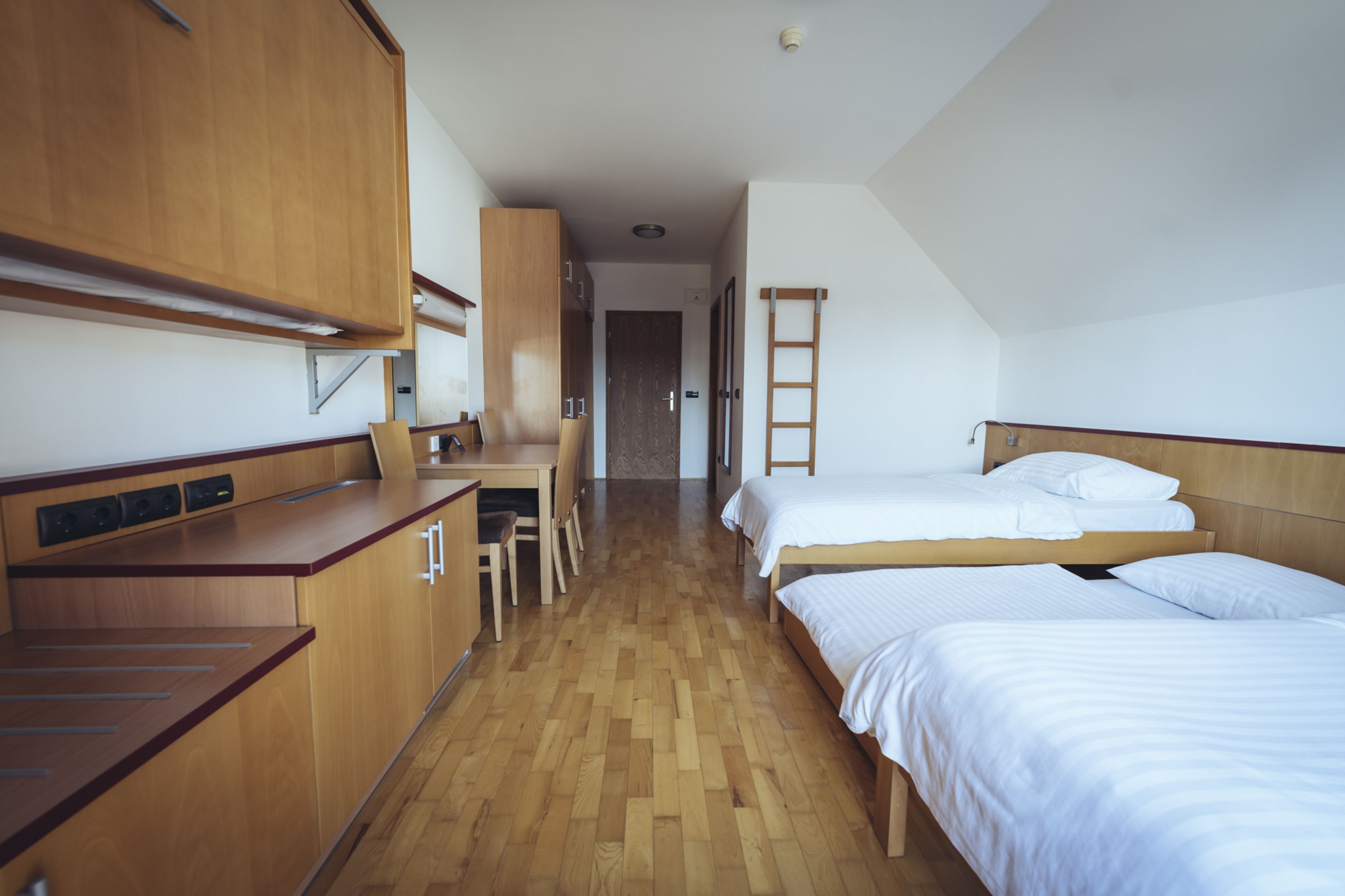 A compact room with a kitchenette, two single beds, a desk, and a chair.