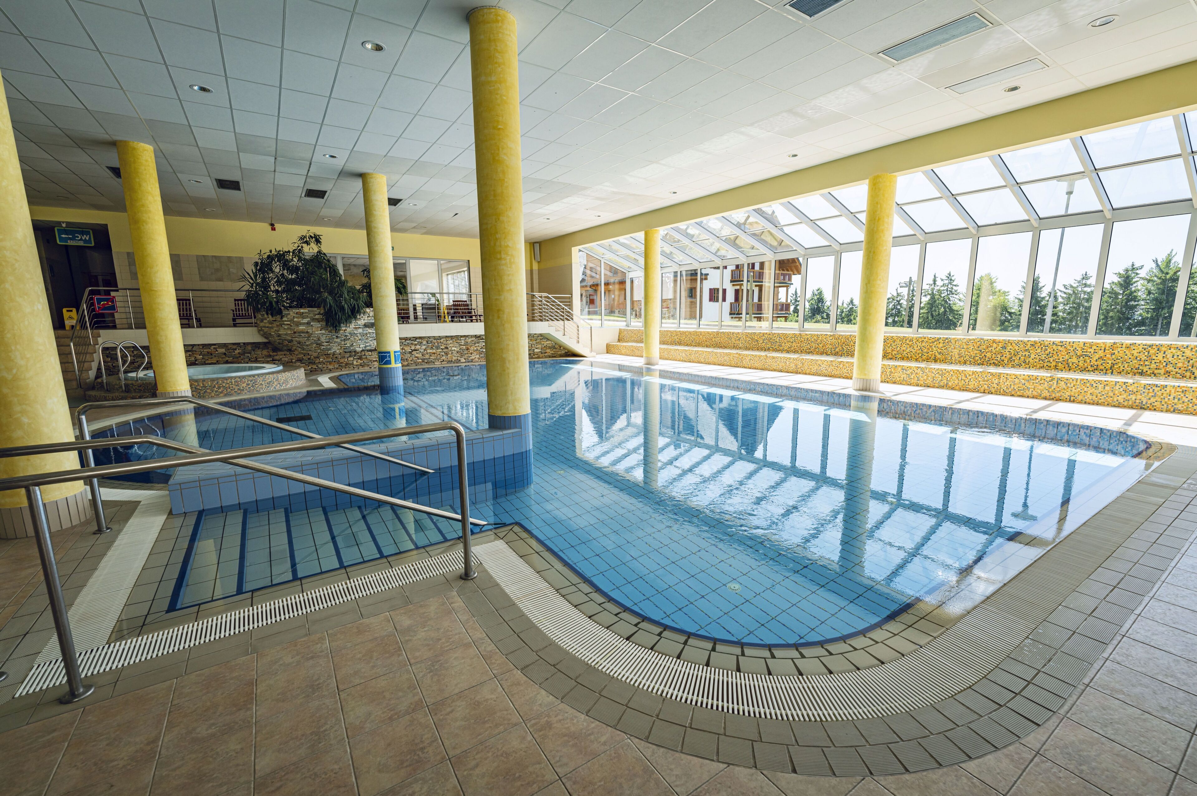 An indoor swimming pool with clear glass walls, yellow pillars, and a tiled floor.