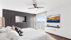 A modern bedroom with a bed, a ceiling fan, a large window, and a wall-mounted TV.