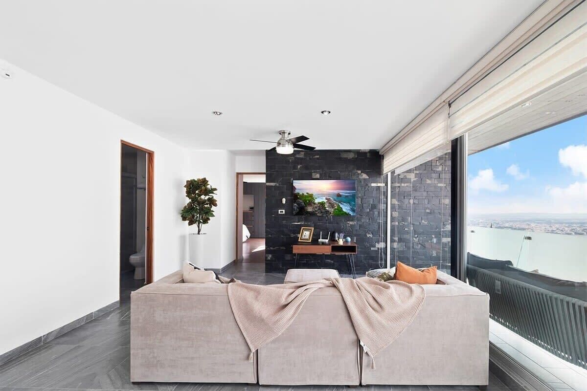 Een moderne woonkamer met een grote bank, een plafondventilator en uitzicht op de stad door grote ramen.