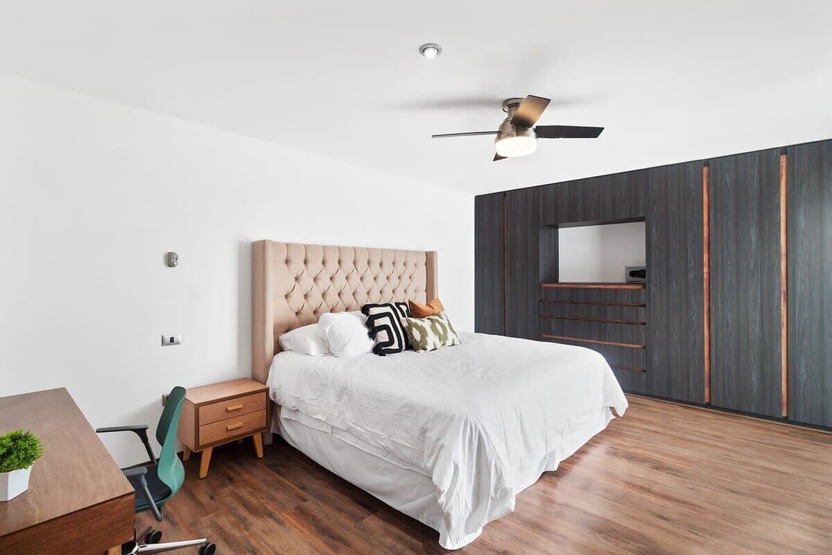 A bedroom with a tufted headboard, a bed with white bedding, a desk with a chair, a small plant, and a ceiling fan.