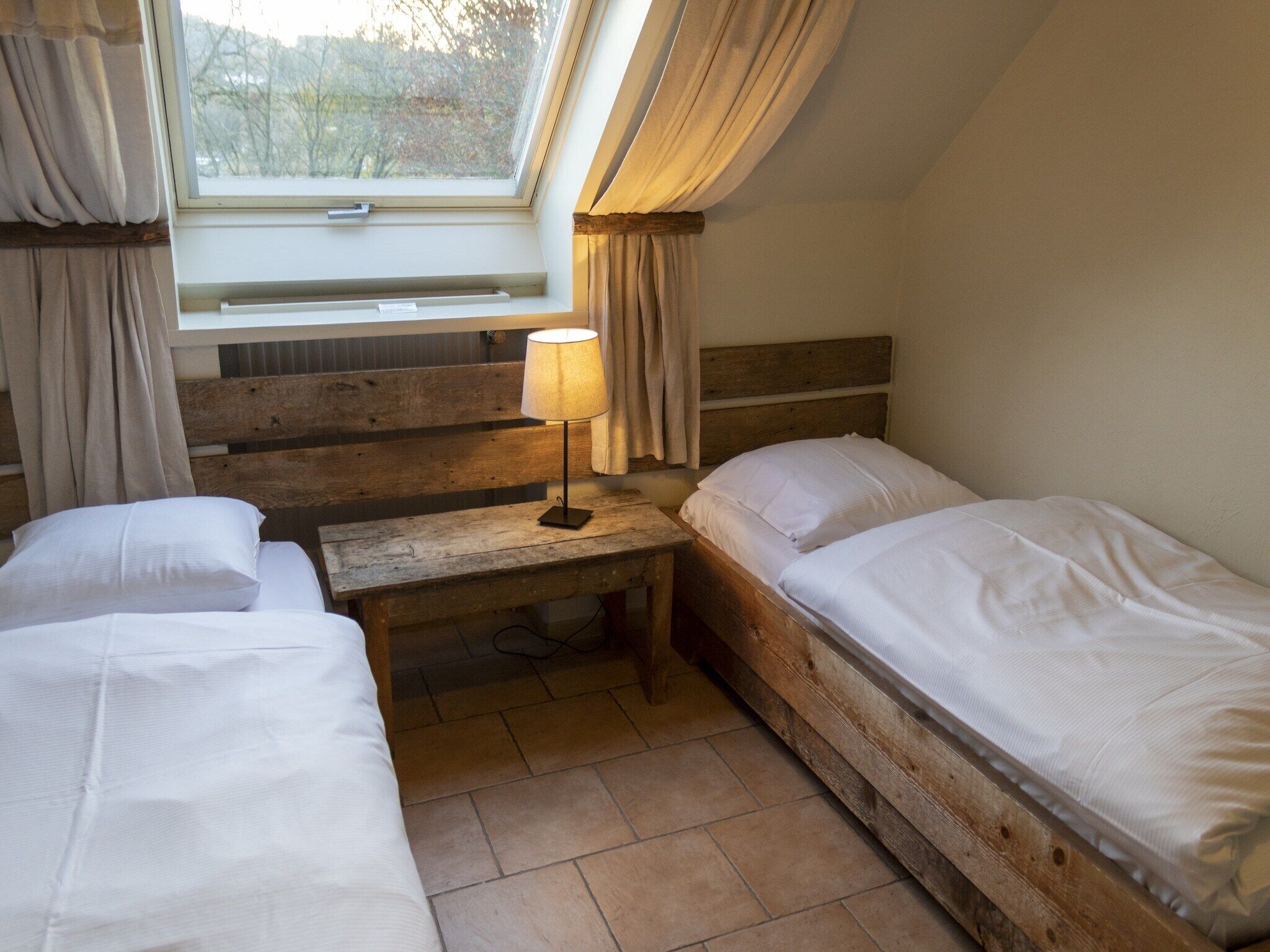 A room with two beds, a wooden bench, a lamp, and a window with curtains.
