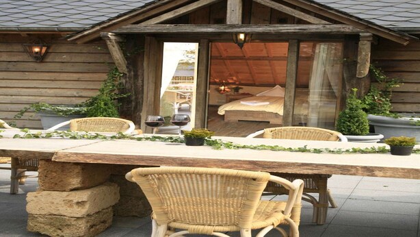 Un bâtiment rustique en bois, doté d’un balcon, de meubles en osier et d’une table dressée pour deux personnes, avec des verres à vin.