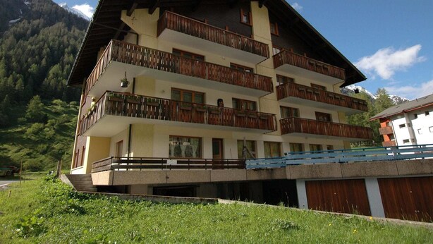 A multi-story building with balconies, set against a mountainous backdrop.
