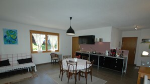 A spacious living area with a dining table, chairs, a sofa, and a kitchen area with a TV and appliances.