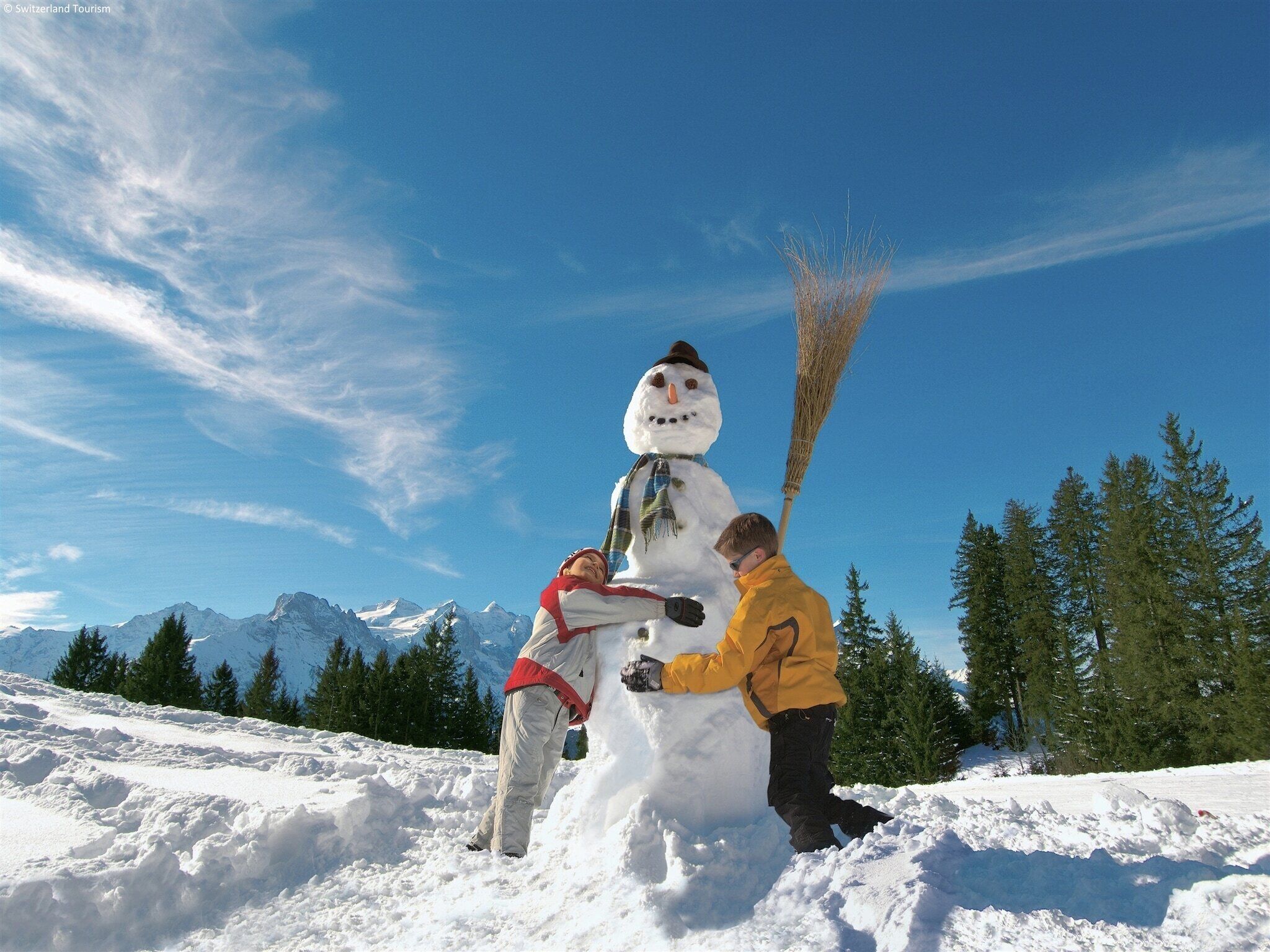 Un bonhomme de neige tenant un balai, et une personne vêtue d’une veste jaune, debout à côté de lui.