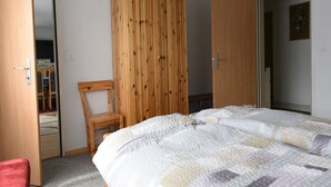 A bedroom with a wooden wardrobe, a bed with a quilt, a chair, and a door leading to another room.