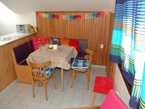 A dining area with a table set for two, wooden chairs, a bench, and a small kitchen area with a microwave.
