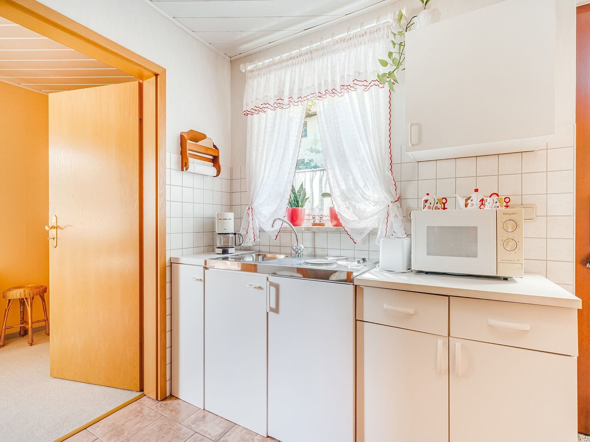 A compact kitchen with white cabinets, a microwave, a coffee maker, and a window with a white curtain.