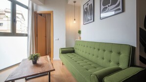 Un sofá verde, una mesa de madera y una planta en maceta en una habitación con una ventana, una puerta y dos cuadros abstractos en la pared.