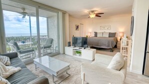 A spacious living area with a balcony, a bedroom with a bed and a TV, and a white coffee table. - Marvelous Views & Steps From The Beach 1608 (Bonita Springs)