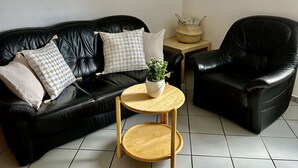 Un sofá de cuero negro con cojines estampados, una mesita de madera con una planta en maceta y un sillón de cuero negro en una habitación con suelo de baldosas.