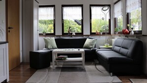 Un salon moderne avec un canapé noir, une table basse blanche et une vue sur la verdure à l’extérieur, grâce à de grandes fenêtres.