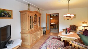 A cozy living room with a wooden cabinet, a television, a dining table with chairs, and a floral ottoman. - Ferienwohnung in Schiltach (Schiltach)
