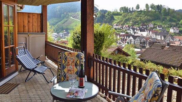 A balcony with two lounge chairs, a table with a bottle and glasses, and a view of a village. - Ferienwohnung in Schiltach (Schiltach)