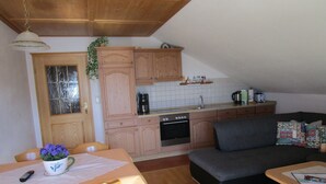 A cozy kitchen with wooden cabinets, a dining table with a vase of flowers, and a dark gray sofa. - Ferienwohnung, Ingenried (Ingenried)