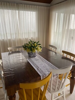Uma mesa de jantar com um vaso de flores amarelas, cadeiras brancas e um jogo americano de renda.