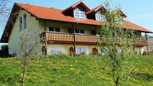 A two-story house with a red-tiled roof, a balcony, and a garden with yellow flowers. - Wohnung in Rotthalmünster mit Balkon (Rotthalmünster)