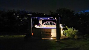 Een barbecueplaats met bakoven en een boogvormige ingang van natuursteen, omringd door een houten pergola met lichtslingers.