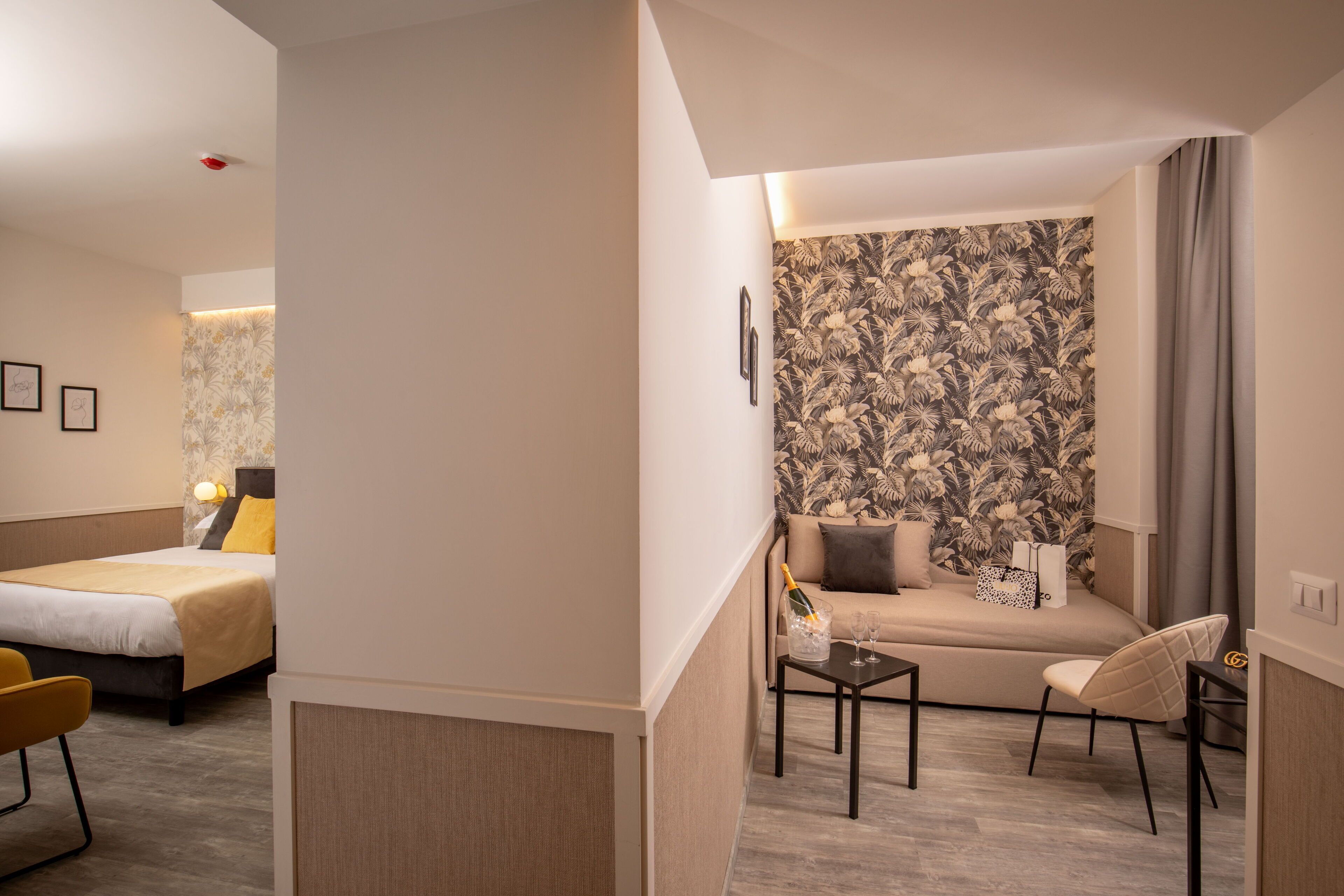 A modern hotel room with a bed, a sofa, a small table with a bottle and glasses, and a chair.