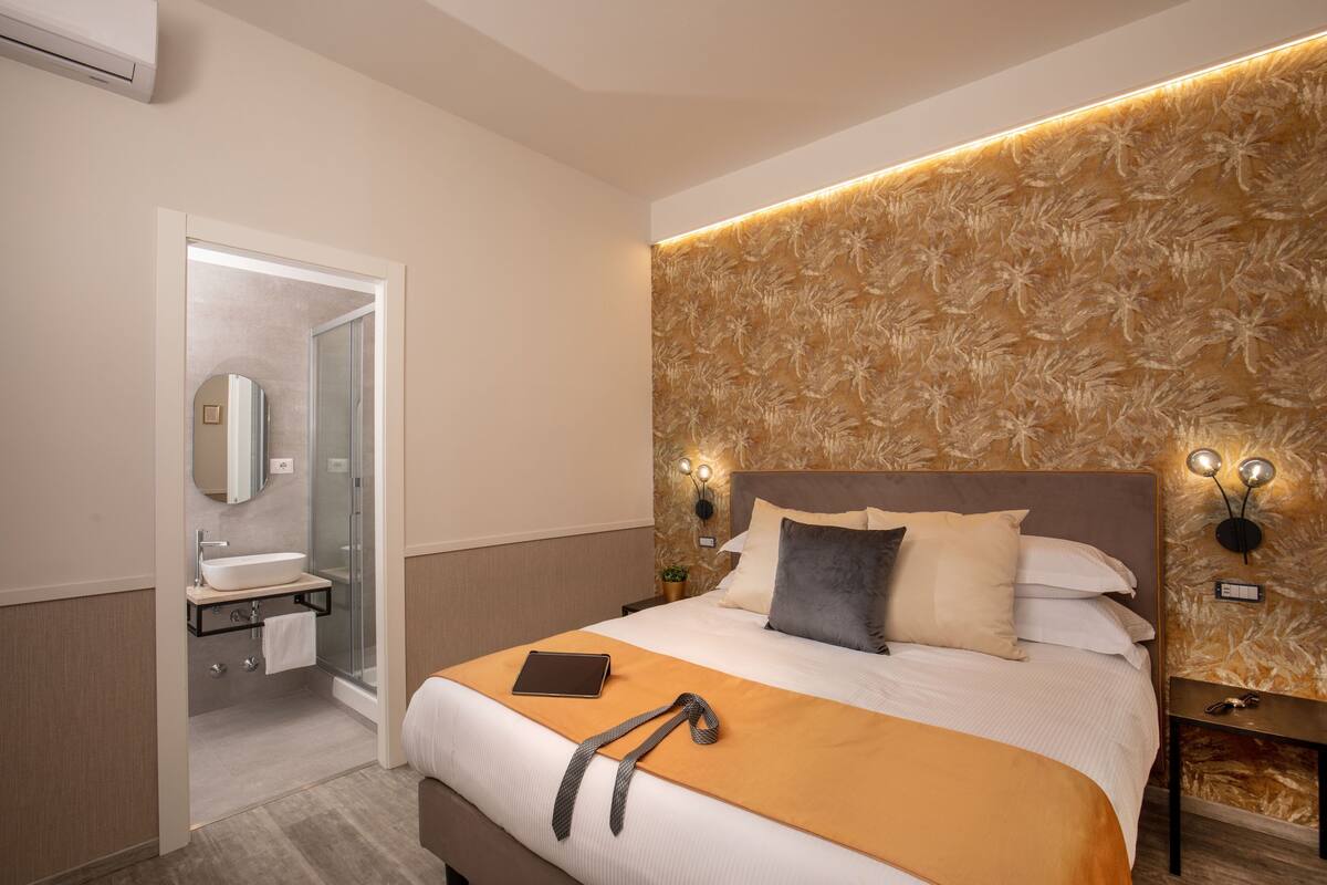 A modern hotel room with a large bed, a bedside table with a lamp, a bathroom with a glass shower, and a decorative wall panel.