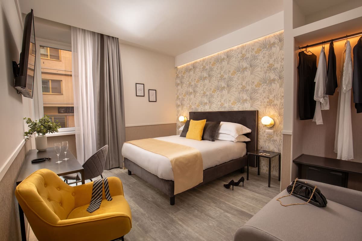 A hotel room with a bed, a desk with a chair, a small table, a coat rack, and a window with curtains.