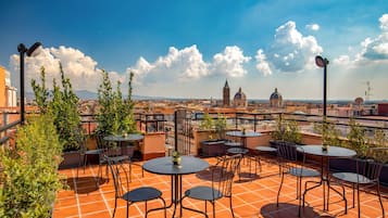 A rooftop terrace with round tables and chairs, potted plants, and a view of a cityscape including a prominent dome and tower.