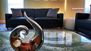 Ein Wohnzimmer mit einer schwarzen Couch, zwei gemusterten Zierkissen, einem Glastisch mit einer Metallskulptur und vier abstrakten Wandbildern.
