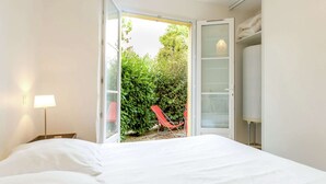 A bedroom with a bed, a nightstand, a lamp, a wardrobe, and an open door leading to a balcony with a chair and greenery.
