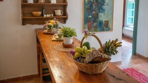 A wooden dining table with a wicker basket containing fruits and a potted plant. A wooden shelf with kitchen items and a painting is in the background.