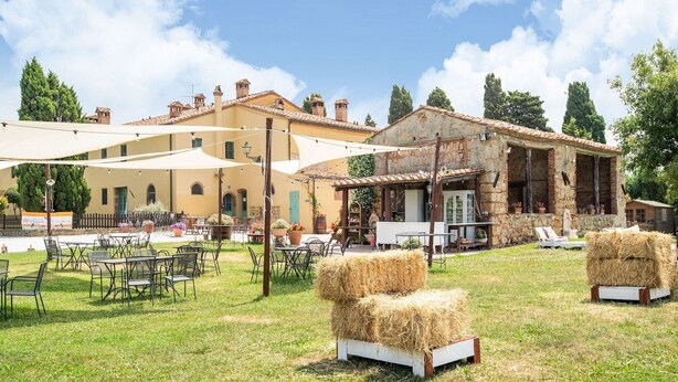 An outdoor seating area with tables and chairs, hay bales, and a building with a stone facade in the background.