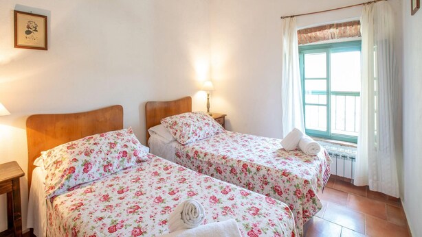 A room with two beds, each with floral bedding, a bedside table with a lamp, a window with curtains, and a framed picture on the wall. - Belvilla by OYO Il Bosco (Collesalvetti (LI))