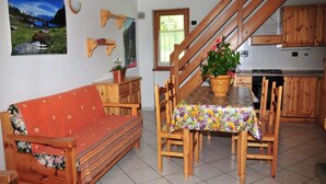 Ein gemütliches Wohnzimmer mit einer Couch, einem Holzespeisetisch und Stühlen. Im Hintergrund ist eine Küche zu sehen.