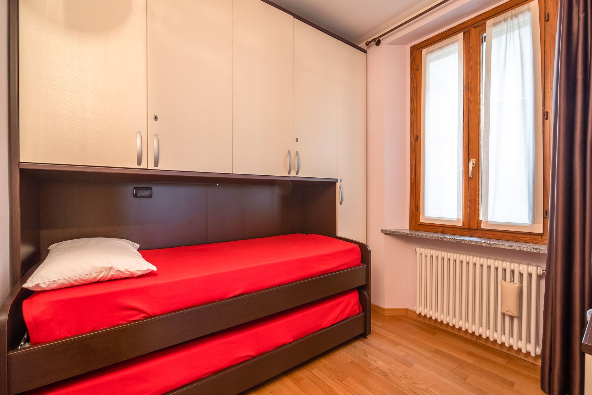 Ein Einzelbett mit roter Matratze in einem Zimmer mit Holzschränken und einem Vorhangfenster.
