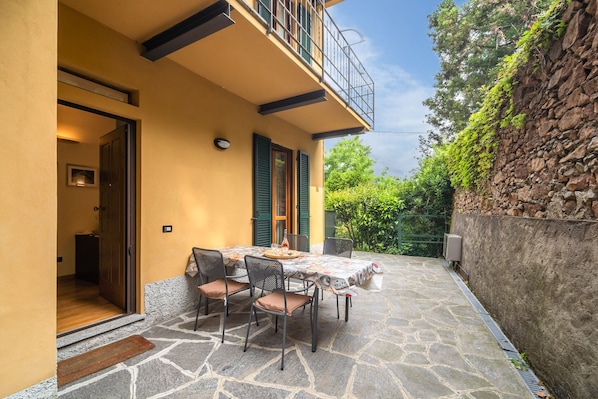 Un patio al aire libre con una mesa dispuesta para comer, una puerta que da a una habitación y un muro de piedra con vegetación.