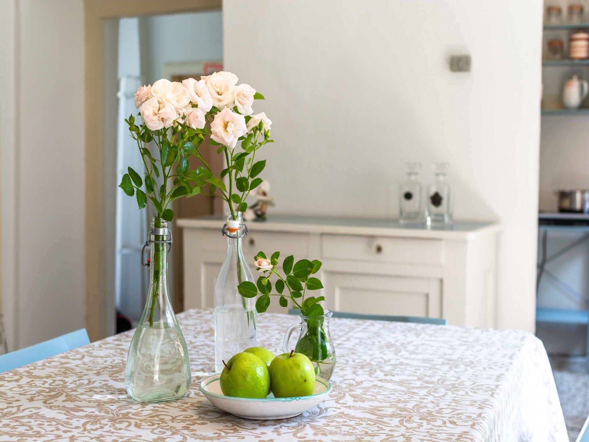 Ein Tisch mit einer Blumenkomposition in Glasflaschen und einer Schale mit grünen Äpfeln.