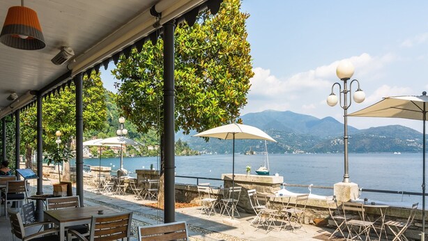 Ein Straßencafé mit Tischen und Stühlen mit Blick auf einen See und Berge im Hintergrund.