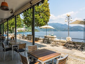 Un café en plein air, avec des tables et des chaises, donnant sur un lac et offrant une vue sur des montagnes en arrière-plan.