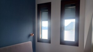 Ein Schlafzimmer mit einem Bett, zwei Fenstern mit Jalousien, einer Wandleuchte und Blick auf ein Gewässer und Berge.