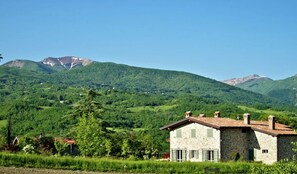 Ein Steinhaus mit geneigtem Dach, umgeben von üppiger Vegetation und Bergen im Hintergrund.