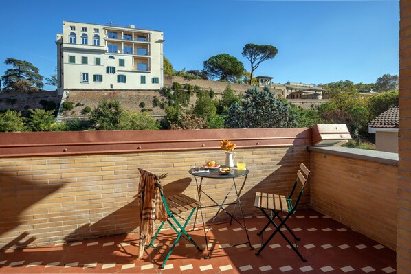 A terrace with a brick wall, two folding chairs, a small round table with a pitcher, plate, and glass of orange juice.