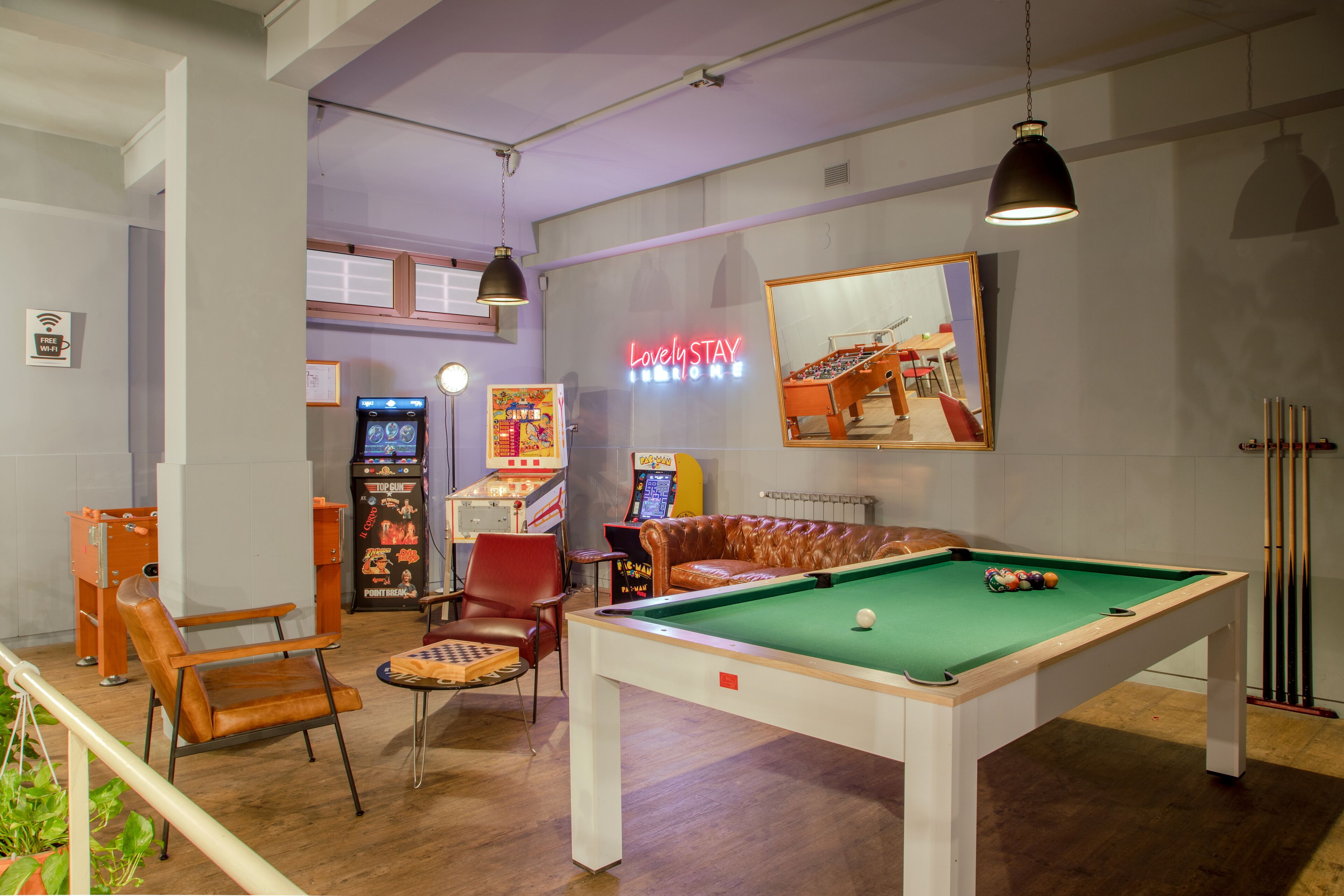 A pool table with a neon sign, a red leather chair, and arcade games in a room with wooden floors.