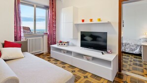 Una habitación de hotel moderna con un televisor grande, un sofá, una mesita, un ventanal con cortinas y vistas a la ciudad.