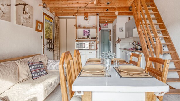 Un rincón comedor acogedor con una mesa de madera dispuesta para dos personas, un sofá con cojines y una escalera que sube a un nivel superior.