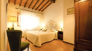 A bedroom with a bed, a chair, a nightstand, a lamp, a wardrobe, and a window with curtains. - Agriturismo Poggio di Montepescoli - Il Ciliegio (Pelago (fi))