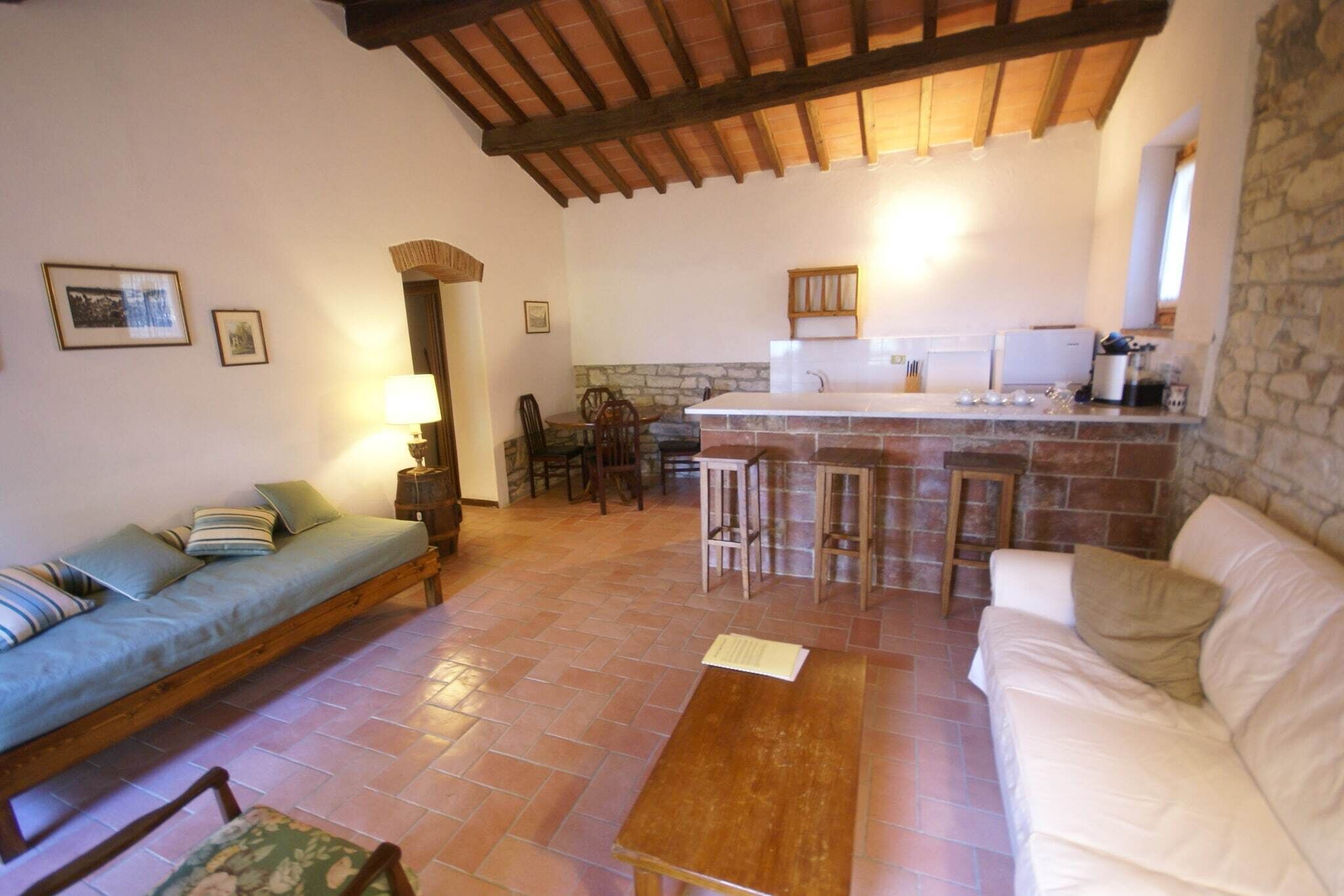 A cozy living room with a sofa, a wooden table, and a kitchen area with a stone wall and wooden beams.