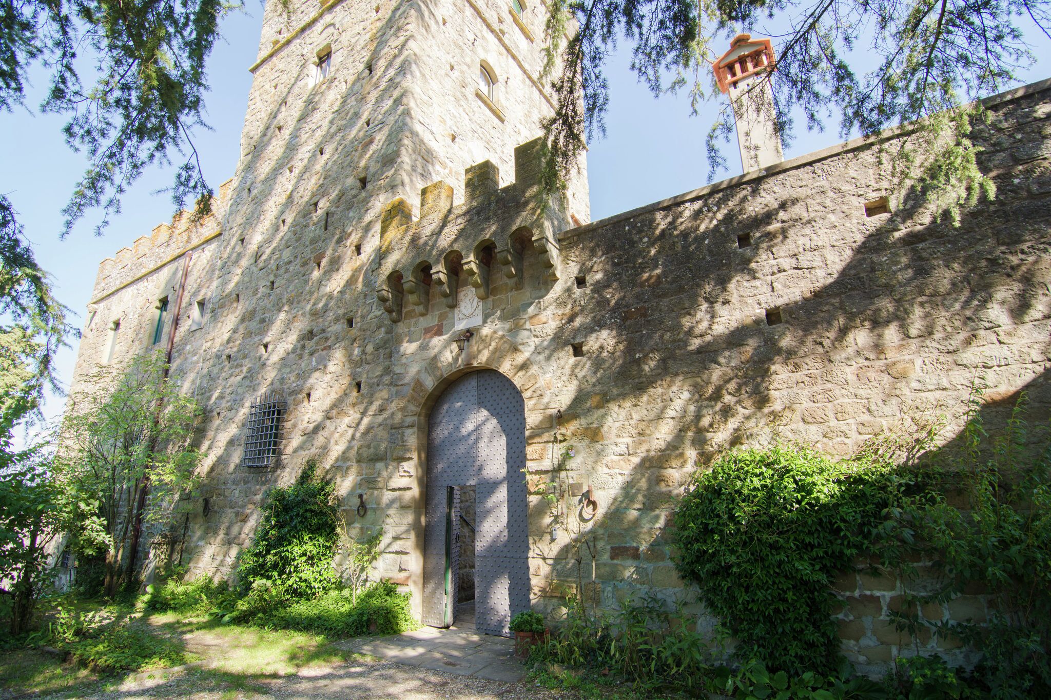 Ein historisches Steinhaus mit einem großen Rundbogenportal, umgeben von Grünpflanzen und blauem Himmel.