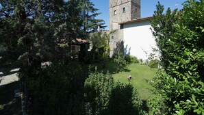 Une tour de pierre historique, surmontée de créneaux, entourée d’une végétation luxuriante et bordée d’un bâtiment blanc au toit recouvert de tuiles.