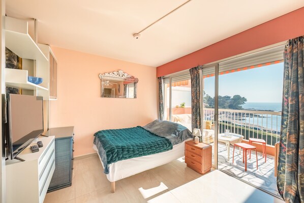 A bedroom with a bed, a TV, a dresser, a mirror, and a balcony with a view of the sea.